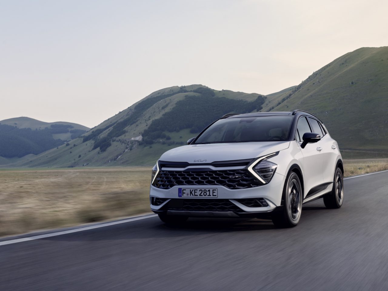 Een witte plug-in hybride SUV rijdt onder een heldere hemel op een verharde weg met groene heuvels op de achtergrond.