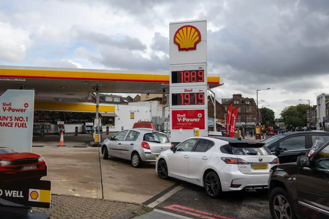 Auto's staan in de rij bij een Shell-tankstation met brandstofprijzen van 188,9 voor loodvrij en 179,9 voor diesel, wat doet denken aan de tarieven voor tanken in Duitsland. Boven het station is een bewolkte hemel zichtbaar.