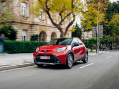 Een rode Toyota Aygo X rijdt door een stadsstraat vol lommerrijke bomen en gebouwen.