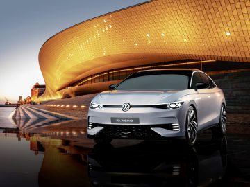 Een strakke, zilveren Volkswagen ID. De Aero-auto staat tijdens de schemering geparkeerd voor een moderne, verlichte architectonische structuur.