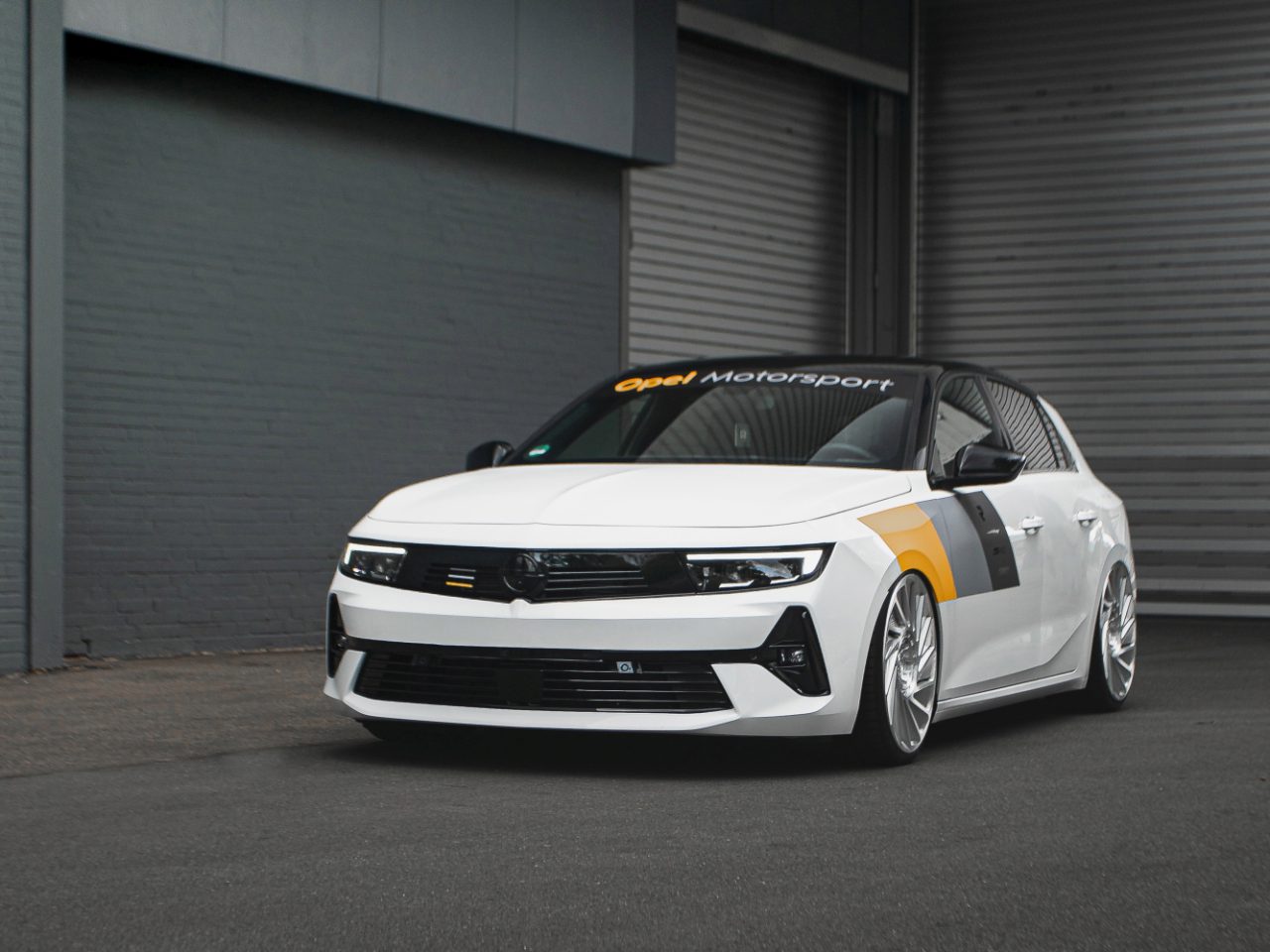 Een witte Opel Astra Motorsport-auto met zwarte en gele details staat geparkeerd op een grijze ondergrond voor een gebouw met industriële roldeuren.