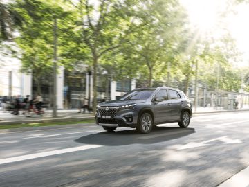 Een grijze Suzuki S-Cross Full Hybrid rijdt door een zonovergoten stadsstraat omzoomd met bomen en gebouwen, met een paar mensen en fietsers op de achtergrond.