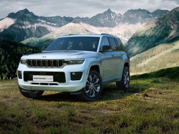 Een witte Jeep Grand Cherokee staat geparkeerd op een grasveld met bergen op de achtergrond. Het voertuig heeft een zichtbaar kenteken aan de voorzijde met de tekst "Grand Cherokee".