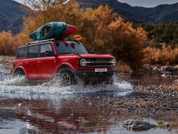 Een rode Ford Bronco SUV met twee kajaks op het imperiaal rijdt door een ondiep beekje, met herfstbomen en bergen op de achtergrond.