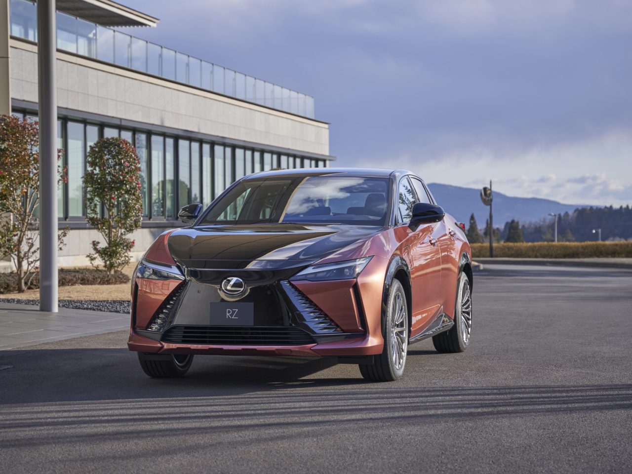 Een gebrand oranje Lexus RZ 450e staat geparkeerd voor een modern gebouw met grote ramen op een verhard terrein, met een bergketen zichtbaar op de achtergrond.