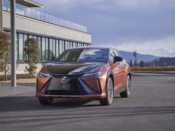 Een gebrand oranje Lexus RZ 450e staat geparkeerd voor een modern gebouw met grote ramen op een verhard terrein, met een bergketen zichtbaar op de achtergrond.