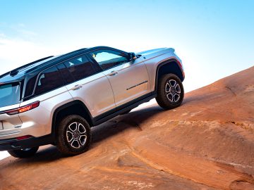 Er wordt een witte Jeep Grand Cherokee getoond die bergop rijdt op een rotsachtig terrein onder een helderblauwe lucht.