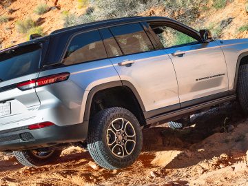 Een zilveren Jeep Grand Cherokee navigeert door een rotsachtig offroad-terrein. Het voertuig is lichtjes gekanteld en de voorwielen zijn hoger, wat zijn capaciteiten en robuuste kenmerken benadrukt.