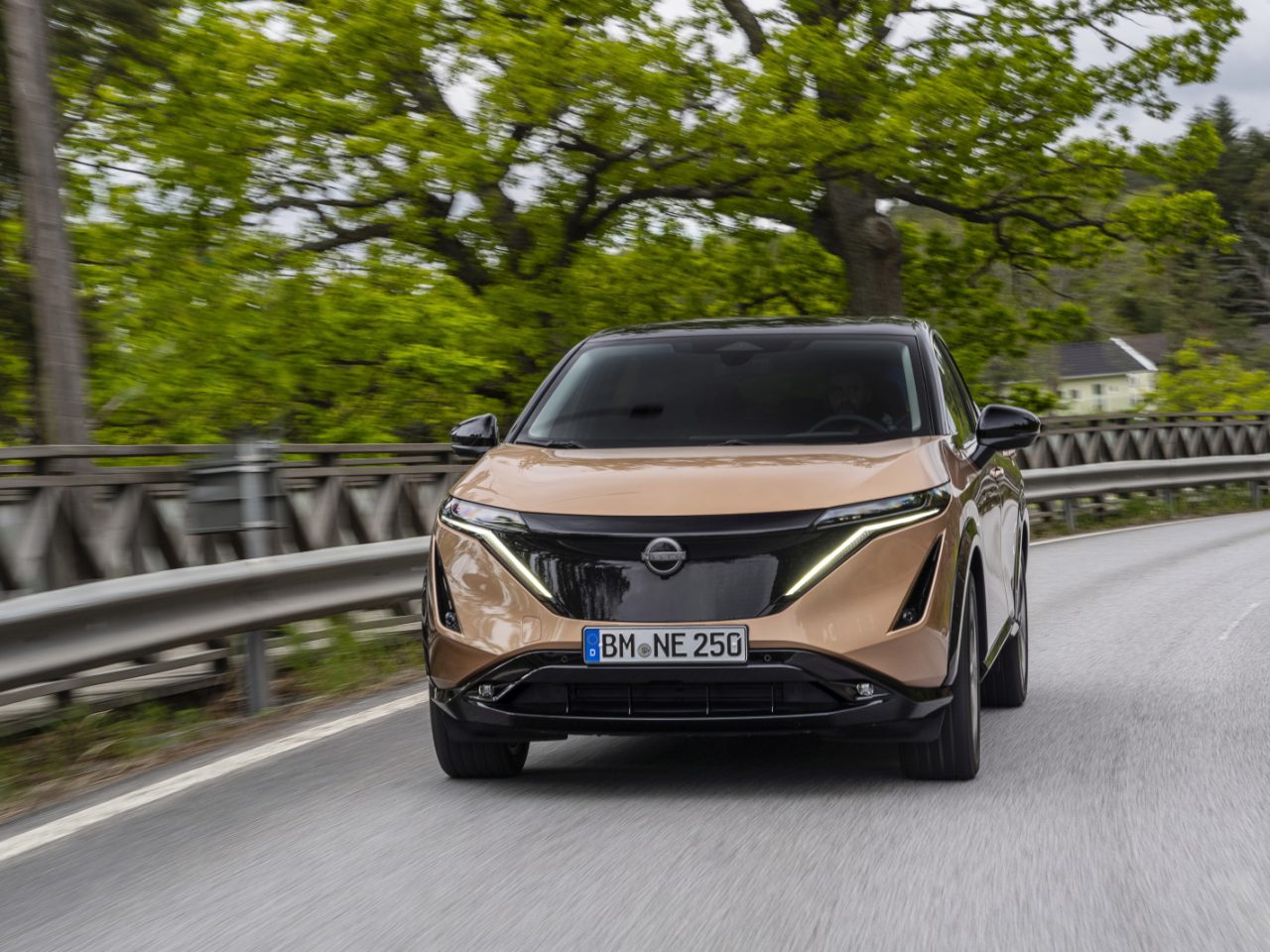 Een lichtbruine Nissan Ariya SUV die over een voorstedelijke weg rijdt met groene bomen op de achtergrond. Op het kenteken van het voertuig staat "BN NE 250.