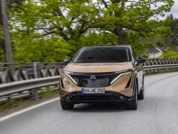 Een lichtbruine Nissan Ariya SUV die over een voorstedelijke weg rijdt met groene bomen op de achtergrond. Op het kenteken van het voertuig staat "BN NE 250.
