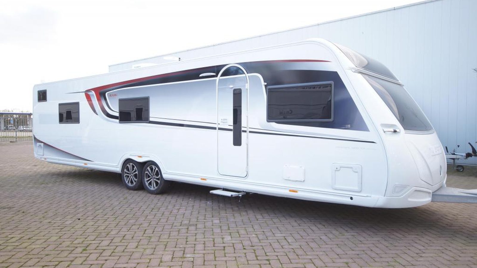 Een grote witte caravan met zwarte en rode accenten, perfect voor wie een caravan wil kopen, staat geparkeerd op een verhard terrein, vastgemaakt aan een voertuig dat niet in het frame staat.