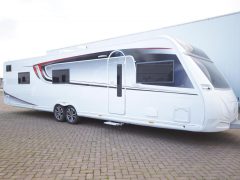 Een grote witte caravan met zwarte en rode accenten, perfect voor wie een caravan wil kopen, staat geparkeerd op een verhard terrein, vastgemaakt aan een voertuig dat niet in het frame staat.