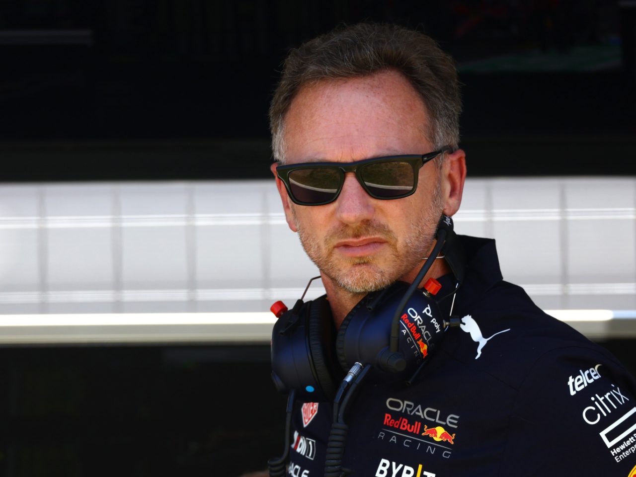 Een man met een zonnebril en een raceteamuniform met een koptelefoon om zijn nek staat voor een garage en lijkt helemaal op het team van Verstappen.