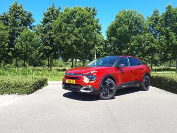 Een rode Citroën C4 staat geparkeerd op een stoep bij groene struiken en bomen onder een helderblauwe lucht. De gestroomlijnde auto heeft zwarte velgen en een geel kenteken.