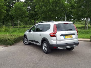 Een slanke zilveren Dacia Jogger staat geparkeerd op een verhard oppervlak, met levendig groen en hoge bomen die een serene achtergrond vormen. Op de achterste kentekenplaat van het voertuig staat "P-515-HF.