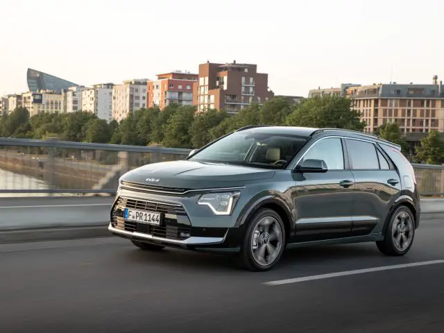 Een grijze Kia Niro EV rijdt over een stadsweg met moderne gebouwen en bomen op de achtergrond en biedt een glimp van innovatie die doet denken aan de rijtest Kia Niro Hybrid-ervaring.