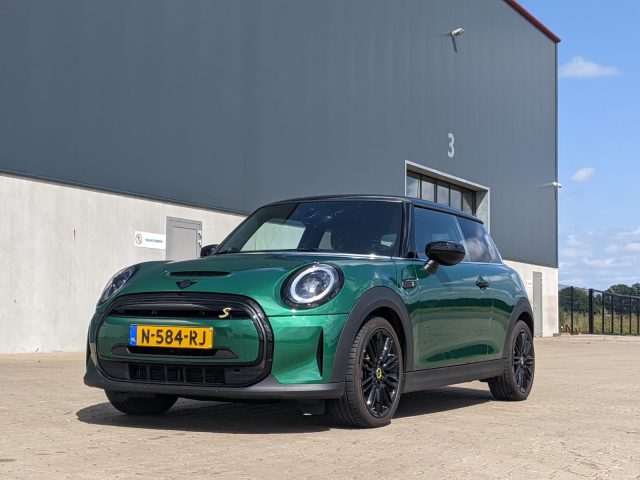 Voor een grijs pakhuis gemarkeerd met het nummer 3 staat een groene elektrische Mini Cooper met Nederlands kenteken geparkeerd.