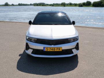 Een witte Opel Astra Sports Tourer met een gele kentekenplaat staat geparkeerd op een verhard oppervlak nabij een watermassa. De achtergrond toont bomen en een heldere hemel.