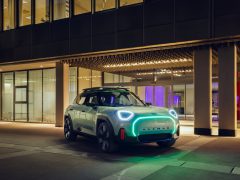Een moderne Mini Aceman SUV met opvallende koplampen en imperiaal staat geparkeerd voor een modern gebouw met grote ramen en 's avonds verlichte interieurs.