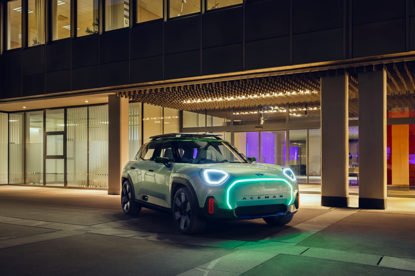 Een moderne Mini Aceman SUV met opvallende koplampen en imperiaal staat geparkeerd voor een modern gebouw met grote ramen en 's avonds verlichte interieurs.