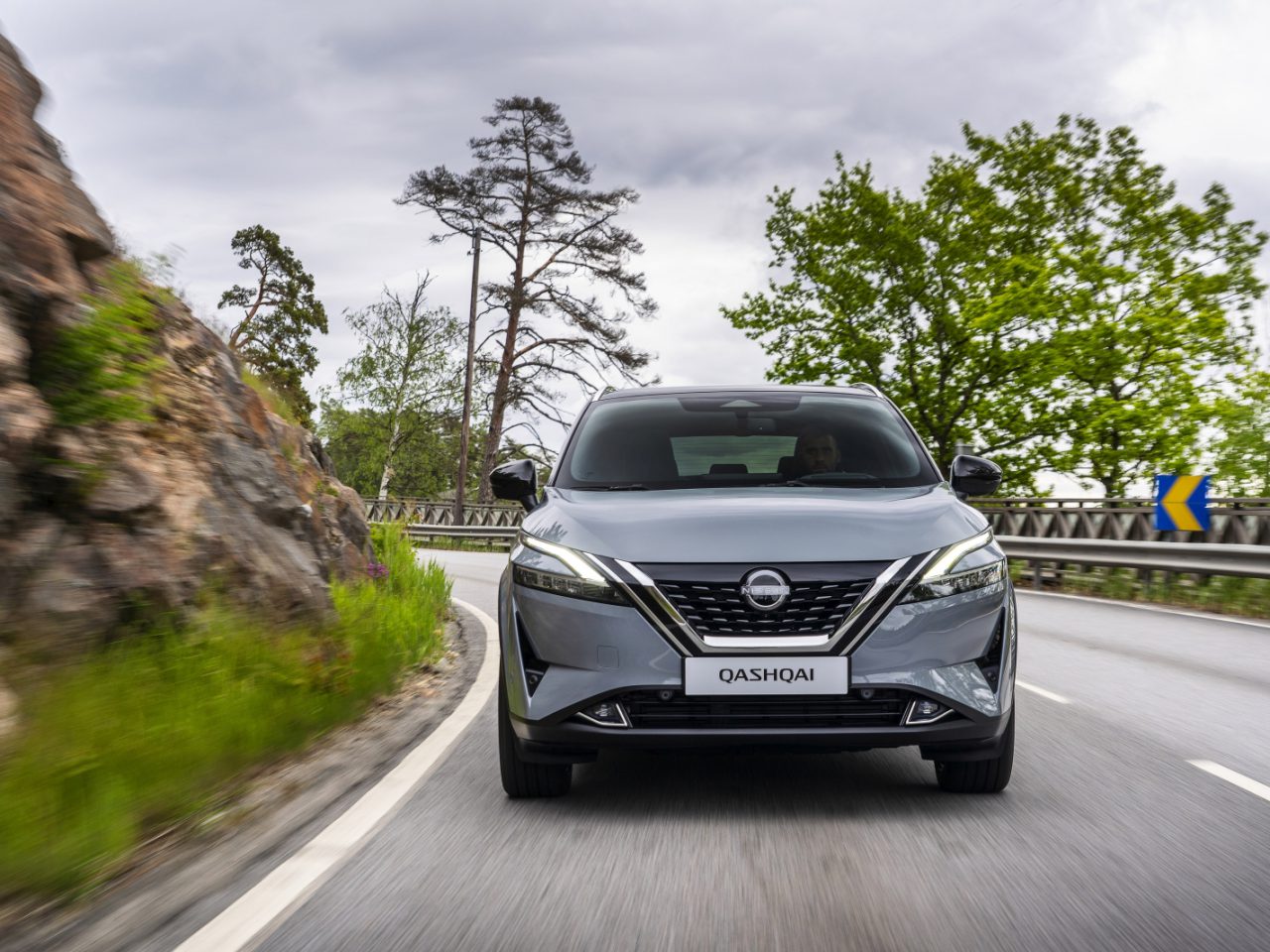 Een grijze Nissan Qashqai e-Power SUV rijdt op een bochtige weg met rotsachtige kliffen aan de linkerkant en bomen aan de rechterkant onder een bewolkte hemel.