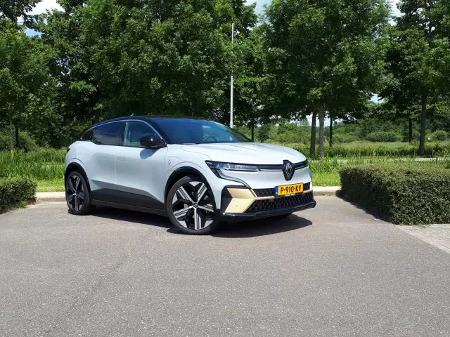 Een zilverkleurige en zwarte Renault Mégane E-Tech Electric met een zichtbare kentekenplaat aan de voorkant staat geparkeerd op een verhard terrein met groene bomen op de achtergrond.