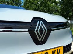 Close-up van de grille van een witte Renault Mégane E-Tech Electric met het Renault-logo, met daaronder een gele kentekenplaat. Bomen en lucht zijn op de achtergrond.