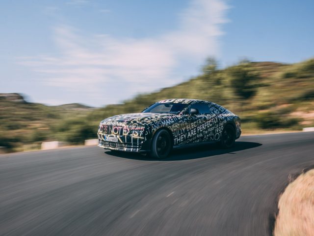Een gecamoufleerde Rolls-Royce Spectre snelt met wazige bewegingen over een bochtige weg, tegen een achtergrond van heuvels en een gedeeltelijk bewolkte lucht.