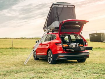Een rode Skoda Enyaq stationwagon met open kofferbak heeft een uitschuifbare kampeerkeuken en een daktent, geparkeerd in een grasveld met in de verte uitzicht op een gebouw met de tekst 'Radar Society'.