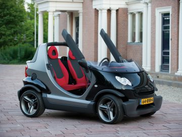 Een kleine, zwart-zilveren cabrio tweezitsauto met een opvallend rood-zwart interieur staat geparkeerd op een verharde oprit voor een gebouw versierd met kolommen.