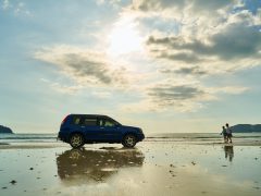 Een blauwe auto staat geparkeerd op een strand in Canada met de oceaan op de achtergrond. Onder een gedeeltelijk bewolkte hemel lopen twee mensen langs de kustlijn.