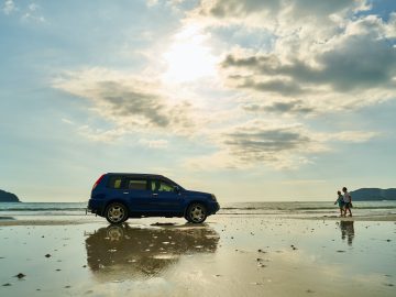 Een blauwe auto staat geparkeerd op een strand in Canada met de oceaan op de achtergrond. Onder een gedeeltelijk bewolkte hemel lopen twee mensen langs de kustlijn.