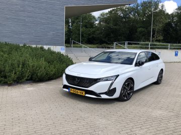 Op een verhard terrein nabij een modern gebouw en groen staat een witte Peugeot 308 met Nederlands kenteken PO-06-HX geparkeerd.