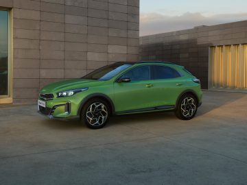 Een groene compacte crossover-SUV Kia XCeed staat geparkeerd voor een modern gebouw met grote glazen ramen en betonnen muren, verlicht door zonlicht.