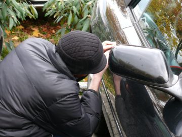 Een persoon in een zwarte jas en muts probeert met gereedschap de deur van wat lijkt op een gestolen auto te ontgrendelen, naast het zwarte voertuig.