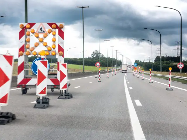 Een snelwegbaan op de A50 is afgesloten met verkeerskegels en slagbomen voor wegwerkzaamheden, onder een bewolkte hemel. Borden geven een snelheidslimiet van 50 km/uur aan en verderop zijn bouwvoertuigen zichtbaar als onderdeel van het onderhoud van de A50.