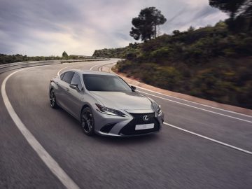 Een slanke Lexus ES sedan rijdt op een gebogen plattelandsweg met bomen en heuvels op de achtergrond onder een bewolkte hemel.