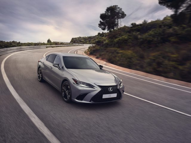 Een slanke Lexus ES sedan rijdt op een gebogen plattelandsweg met bomen en heuvels op de achtergrond onder een bewolkte hemel.