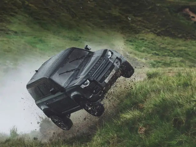Een zwart terreinvoertuig vliegt door de lucht terwijl het een met gras begroeide heuvel oprijdt, wat een scène rechtstreeks uit een James Bond-film oproept, met een ander voertuig gedeeltelijk zichtbaar op de achtergrond.