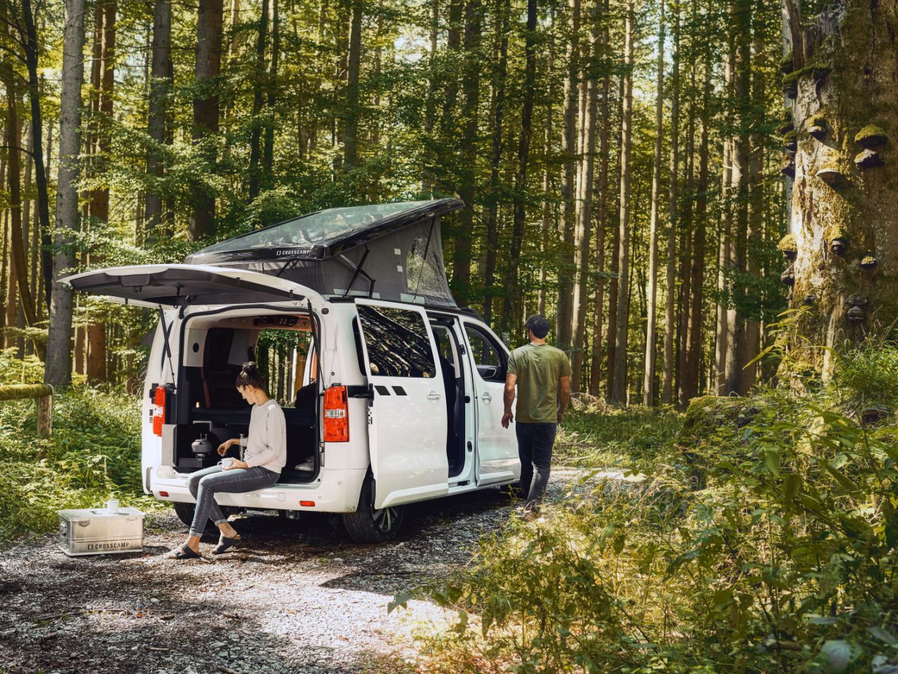 Een wit busje, ingericht als camper met een uitzettentdak, staat in een bos geparkeerd met de achterdeur open. Eén persoon zit op de bumper van het busje en werkt op een laptop, terwijl een ander wegloopt van het voertuig.