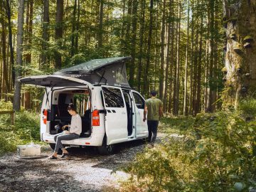 Een wit busje, ingericht als camper met een uitzettentdak, staat in een bos geparkeerd met de achterdeur open. Eén persoon zit op de bumper van het busje en werkt op een laptop, terwijl een ander wegloopt van het voertuig.