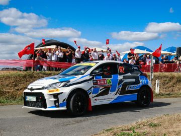 Een wit-blauwe Yaris-raceauto met het nummer H2 op de zijkant snelt tijdens een race over de weg, terwijl op de achtergrond een menigte toeschouwers met rode vlaggen juicht.