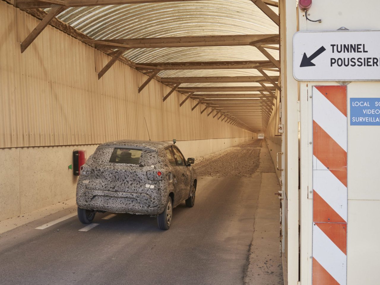Een stoffige Renault rijdt door een lange, afgesloten tunnel met het opschrift 'Tunnel Poussieres'. Borden wijzen op videobewaking in de omgeving.