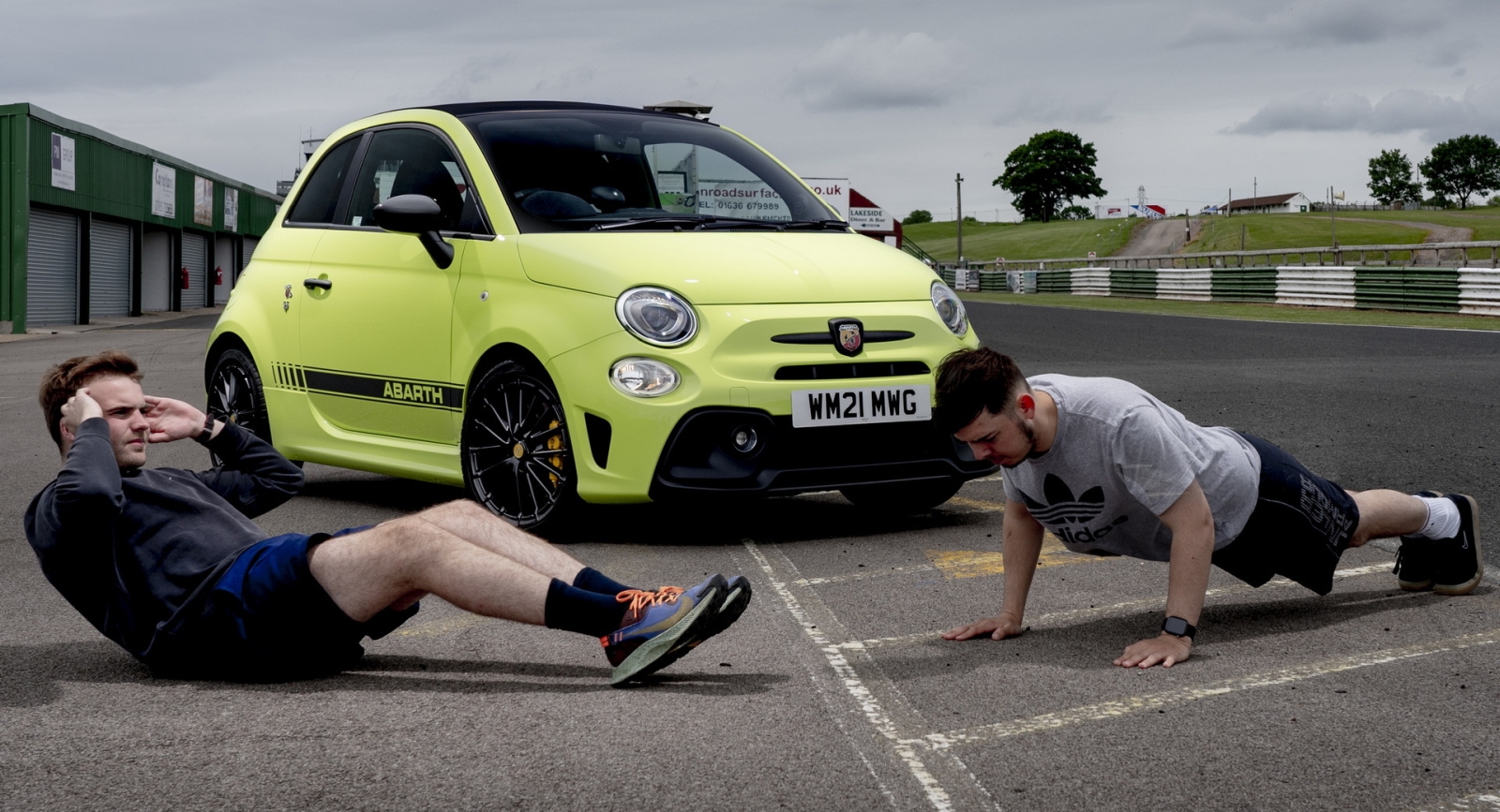 Twee mannen sporten op de grond voor een geparkeerde gele Abarth 595 op een circuit. De ene man doet sit-ups, terwijl de ander push-ups doet.