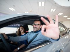 Een man bestuurt een auto en maakt een 'OK'-gebaar met zijn hand. Een vrouw zit glimlachend op de passagiersstoel. Ze zitten allebei in een goed verlichte ruimte, mogelijk bij een autodealer, en bespreken wanneer alle risico's de beste optie kunnen zijn voor hun nieuwe auto.