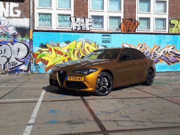 Een gouden Alfa Romeo Giulia Veloce staat geparkeerd voor een kleurrijke, met graffiti bedekte muur, met daarboven meerdere grote ramen.