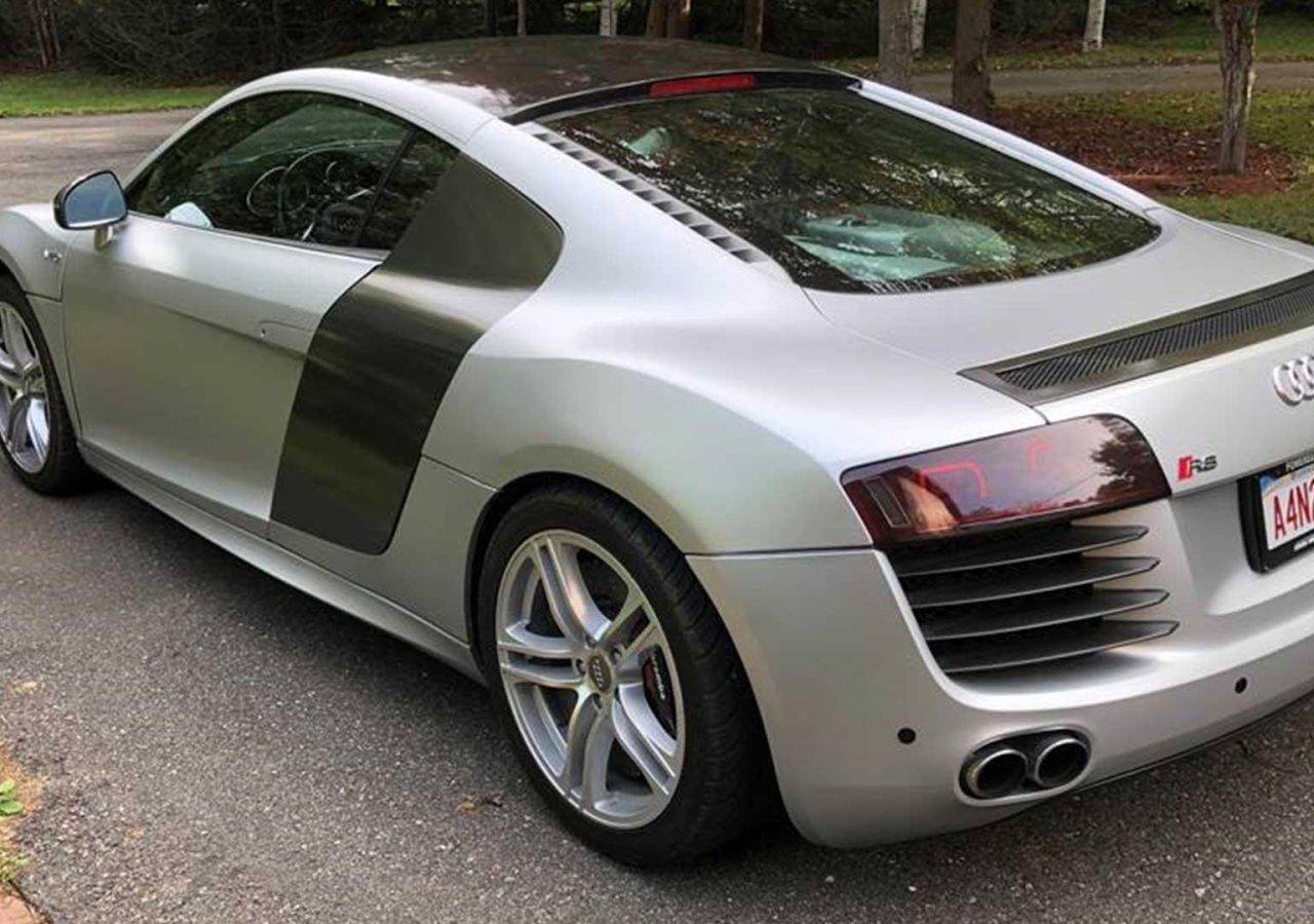 Op de oprit staat een slanke zilveren Audi R8-sportwagen geparkeerd, vastgelegd vanuit een hoek linksachter, met trots zijn "LANCER8"-kentekenplaat.
