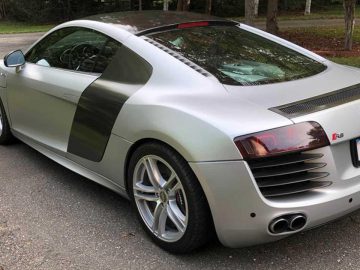 Op de oprit staat een slanke zilveren Audi R8-sportwagen geparkeerd, vastgelegd vanuit een hoek linksachter, met trots zijn "LANCER8"-kentekenplaat.