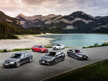 Een groep van vijf auto's, waaronder een strakke Audi RS 6, staat geparkeerd op een weg bij een rustig meer met majestueuze bergen op de achtergrond.