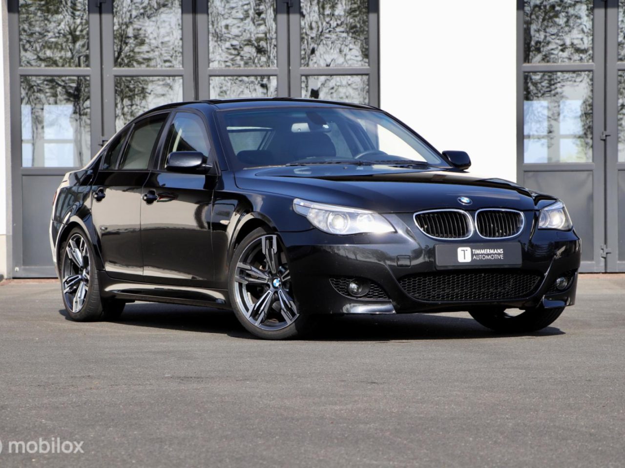 Een zwarte BMW 530i sedan staat geparkeerd op een verhard oppervlak voor een gebouw met grote glazen ramen en grijze deuren.